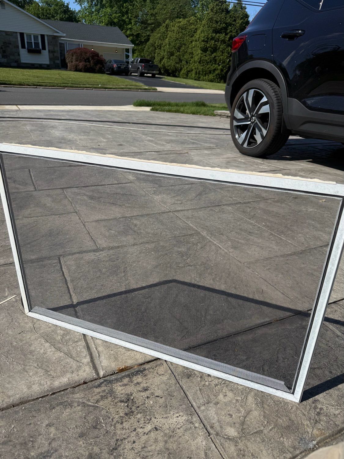 A white-framed rectangular window screen leans against a concrete driveway in a suburban neighborhood.