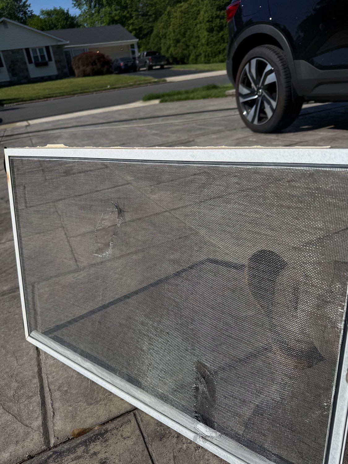 A white-framed window screen with a visible tear, sitting on a concrete driveway with a car partially visible in the back.