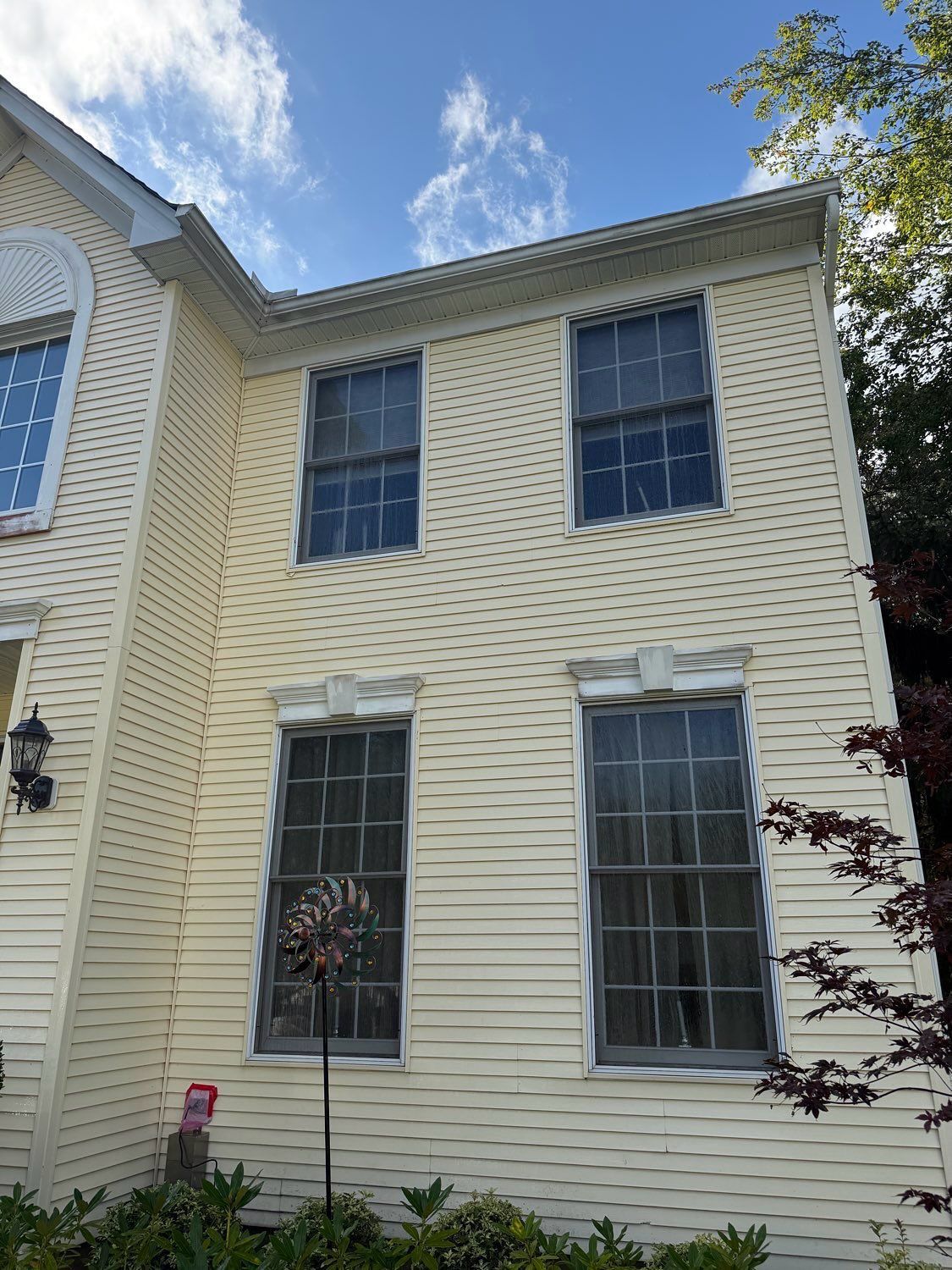 A two-story light yellow house exterior with white trim, tall windows, and green landscaping in the foreground.