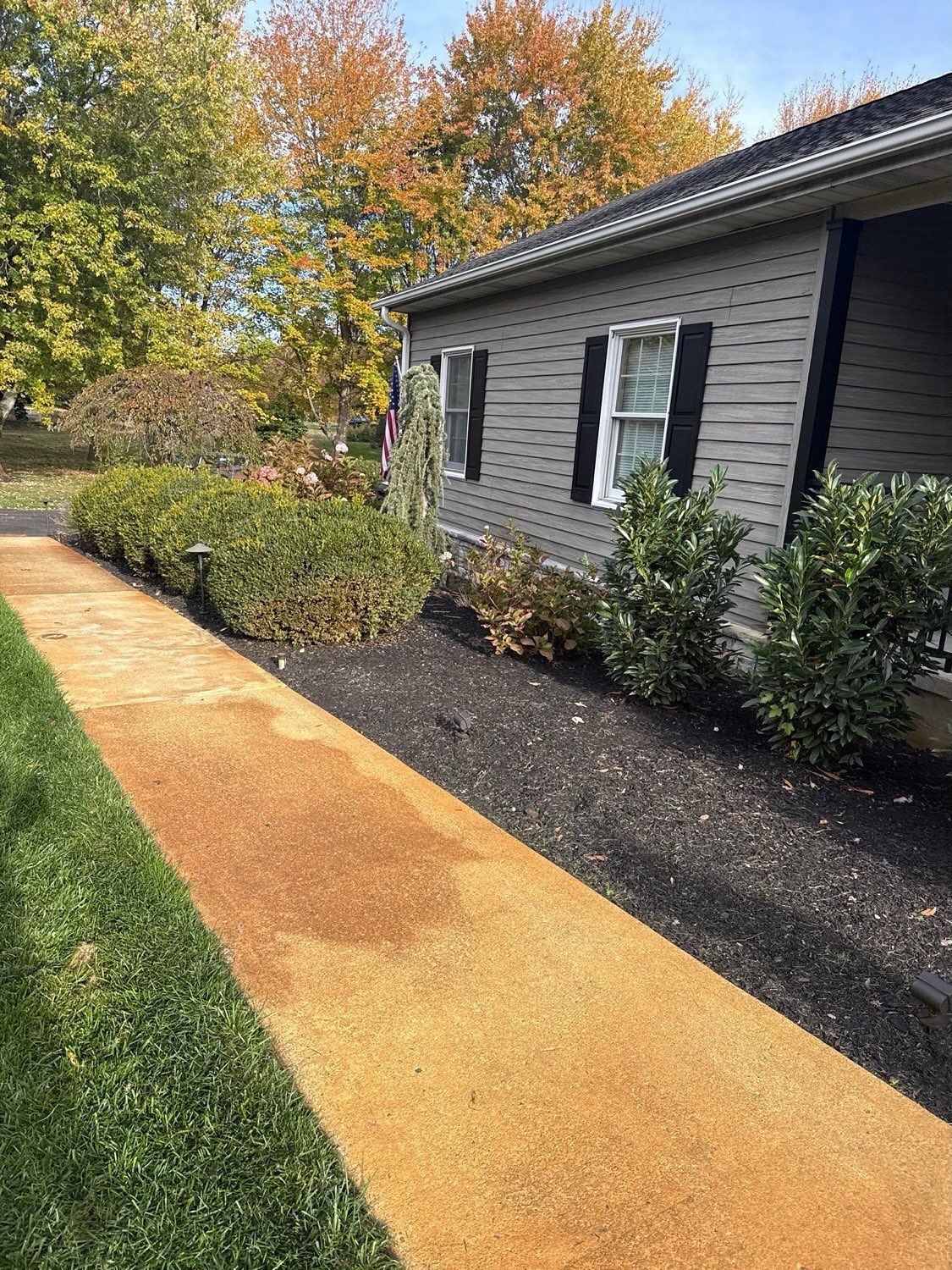 A beige concrete walkway leads past a house with grey siding, black shutters, and landscaping, against autumn trees.