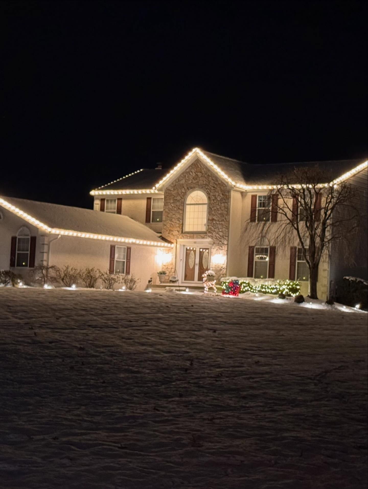 A two-story house at night covered in snow, decorated with bright white string lights along the roofline and landscaping.
