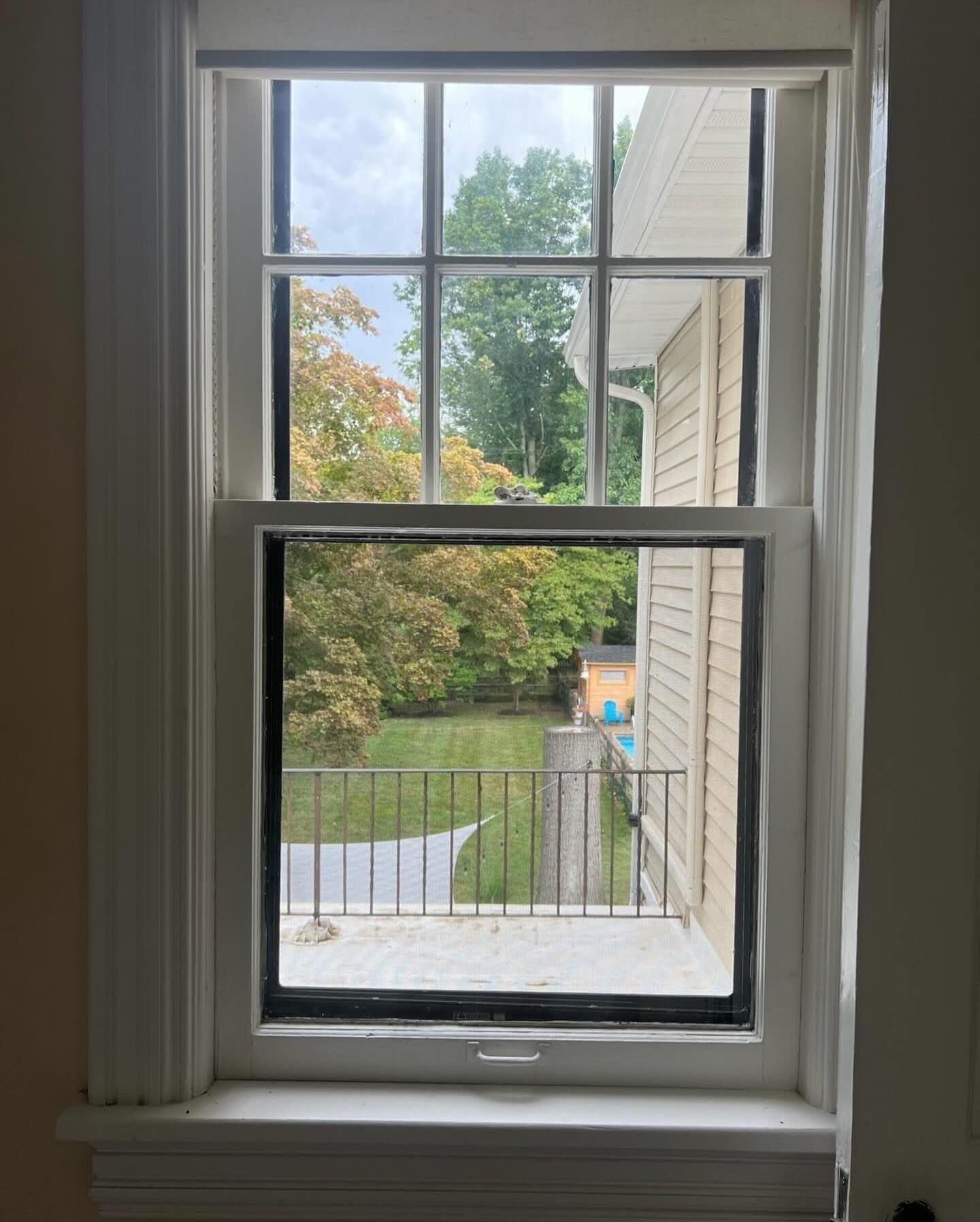 A view through a white-framed, double-hung window looking out at a grassy backyard, trees, and a neighboring house.