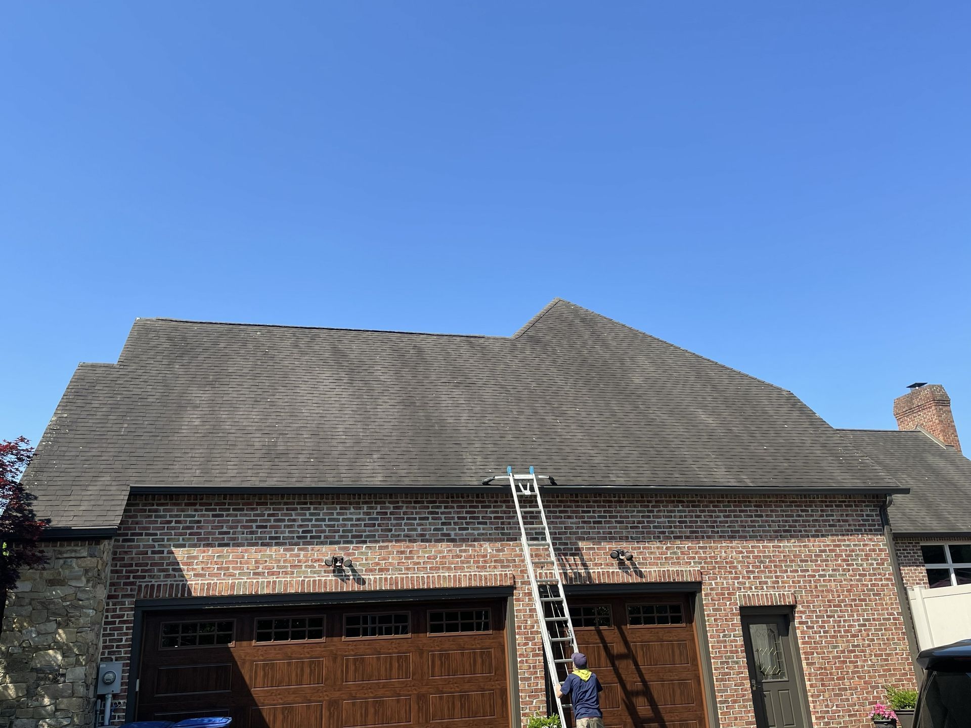A person on a ladder working on the roof of a brick house with a multi-gabled shingle roof under a clear blue sky.