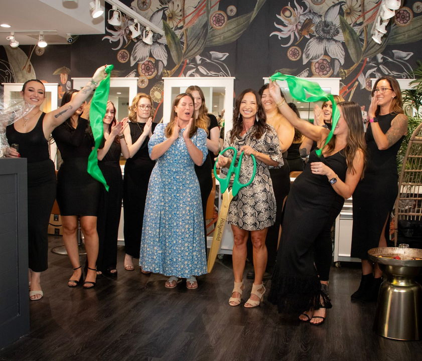 Group of women celebrating inside a boutique, waving green fabric. Some clap. Floral wallpaper in background.