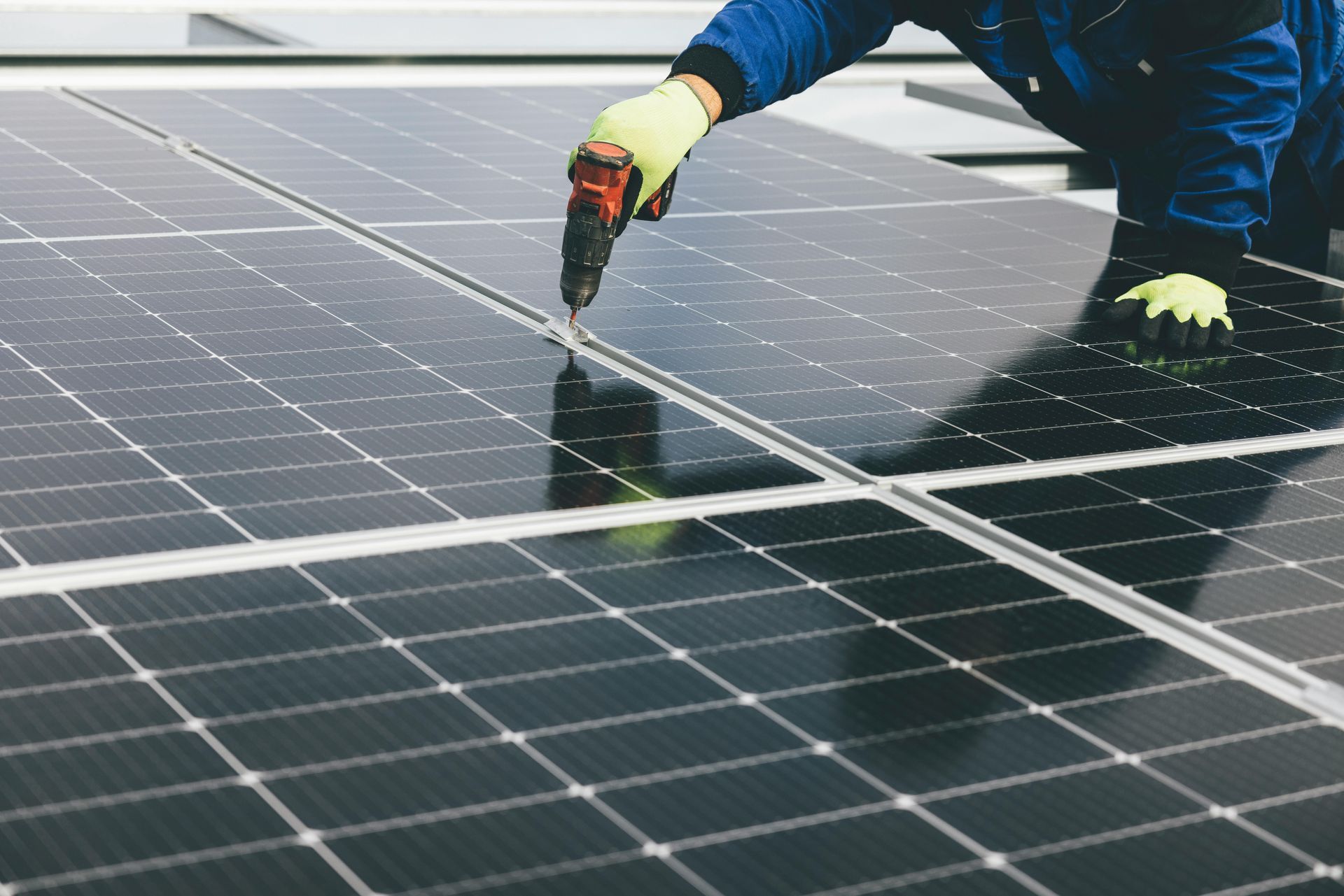 Solar panel installer uses a tool, wearing gloves and blue work clothes, on a rooftop, connecting solar panels.