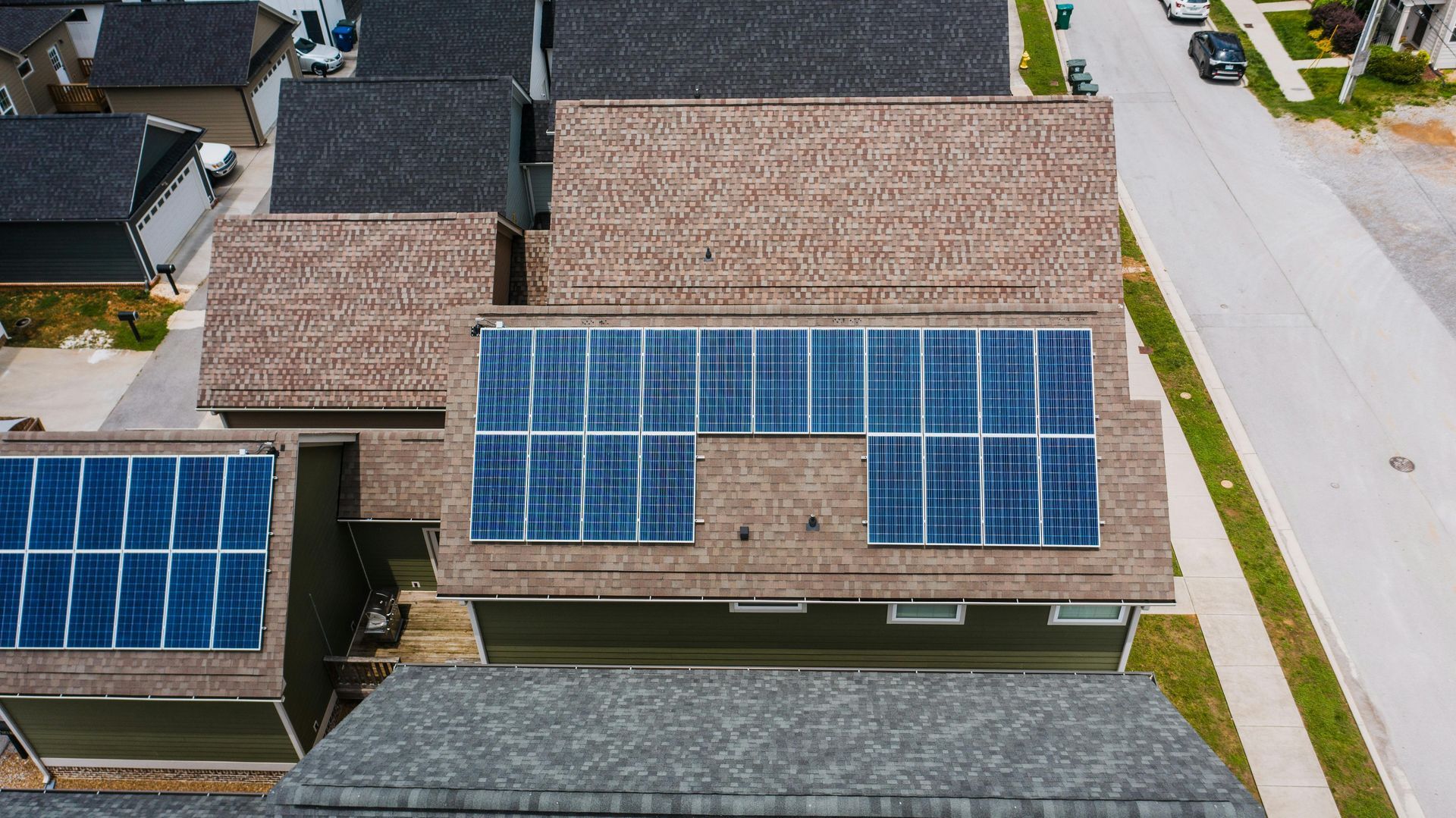 Overhead view of rooftops with solar panels installed on a brown shingled roof, in a residential neighborhood.