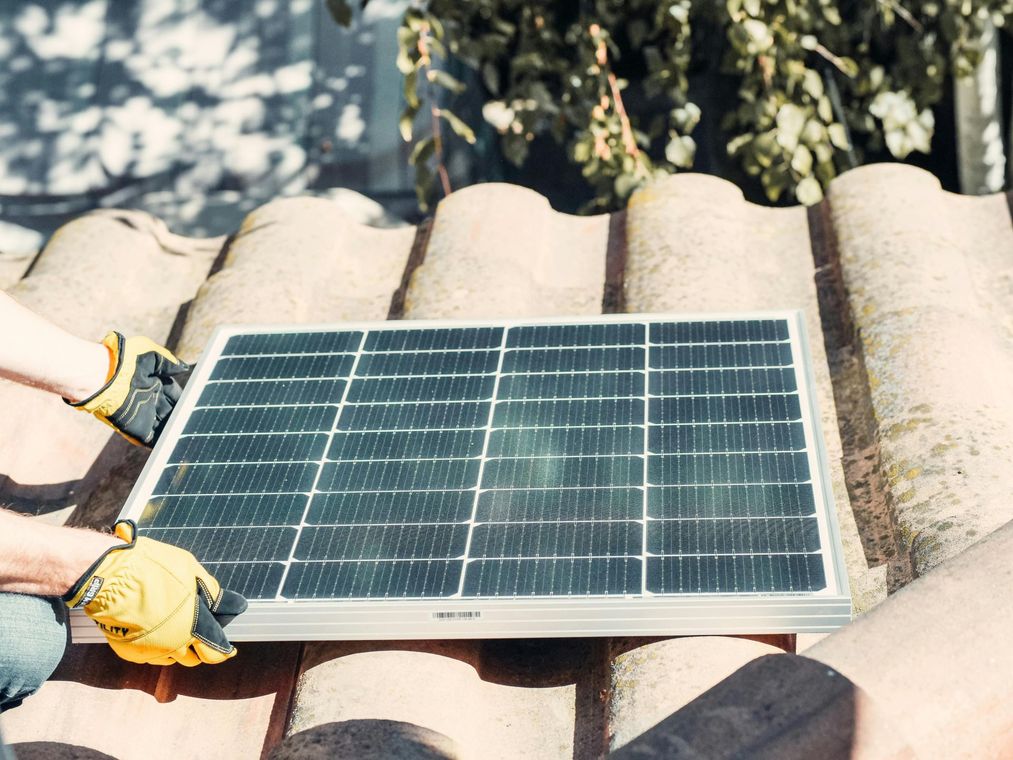Two hands in yellow gloves placing a solar panel on a tiled roof.