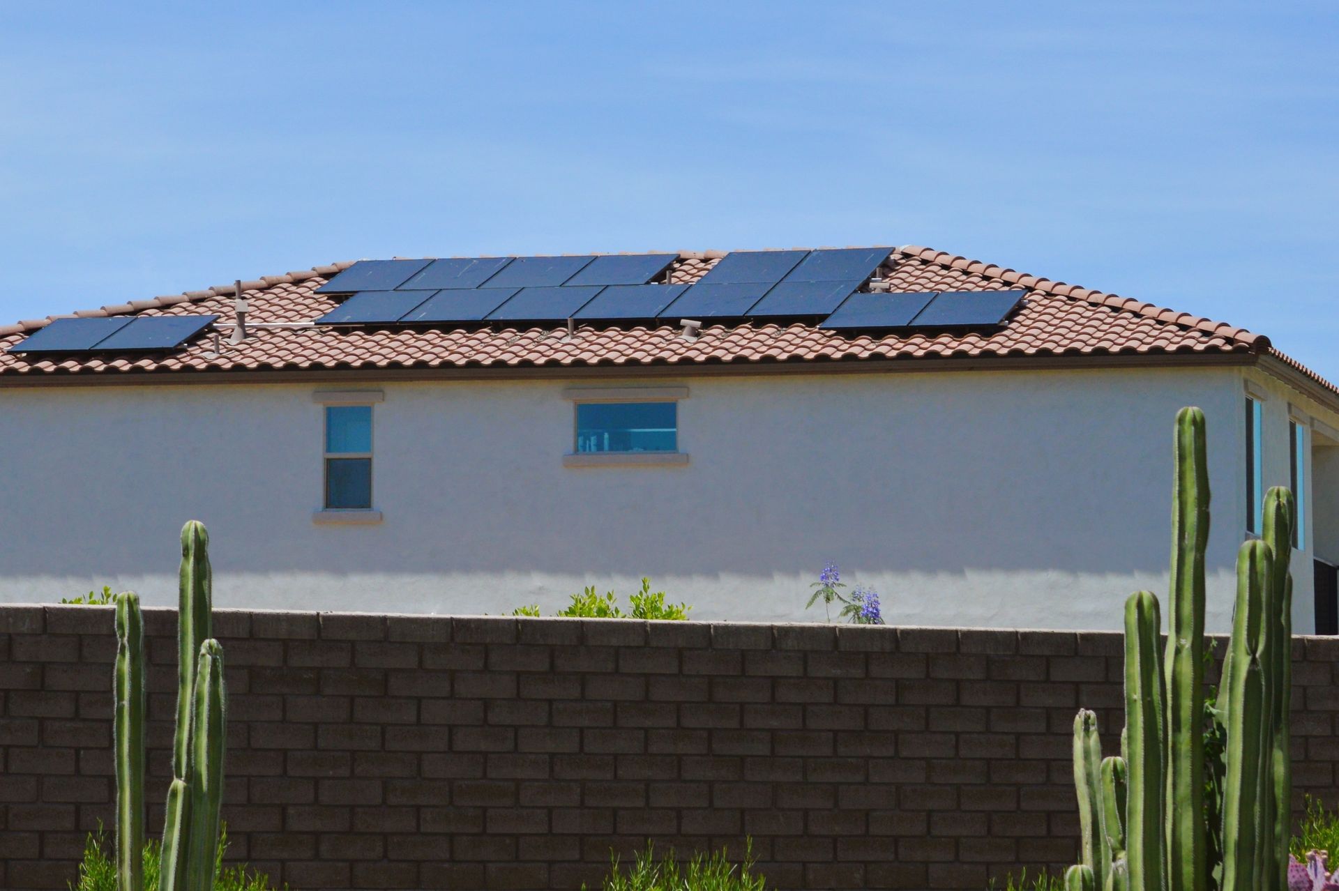 Solar panels on a terracotta-tiled roof of a white house, with a brick wall and cactus in the foreground under a blue sky.