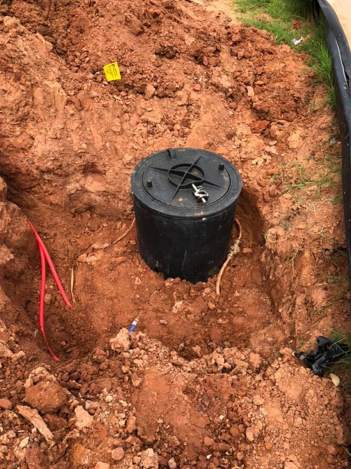 Black utility access box in a dirt hole with red and white wires extending.