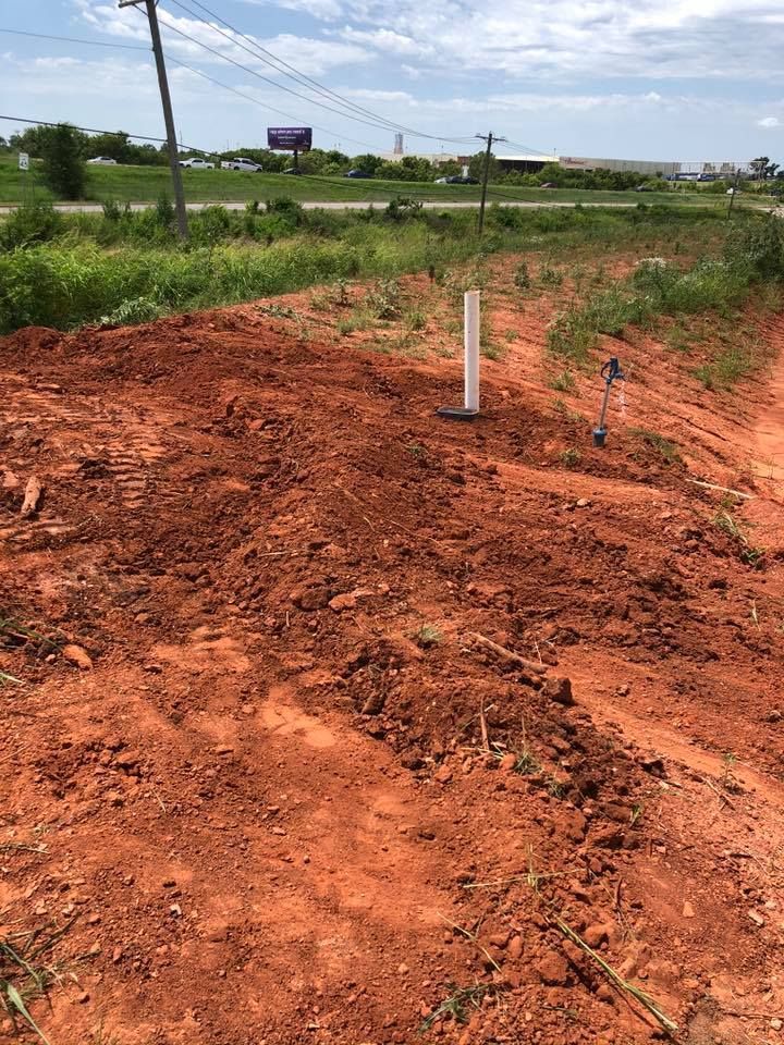 Red dirt plot with white pipe and blue valve; grass and road in background.