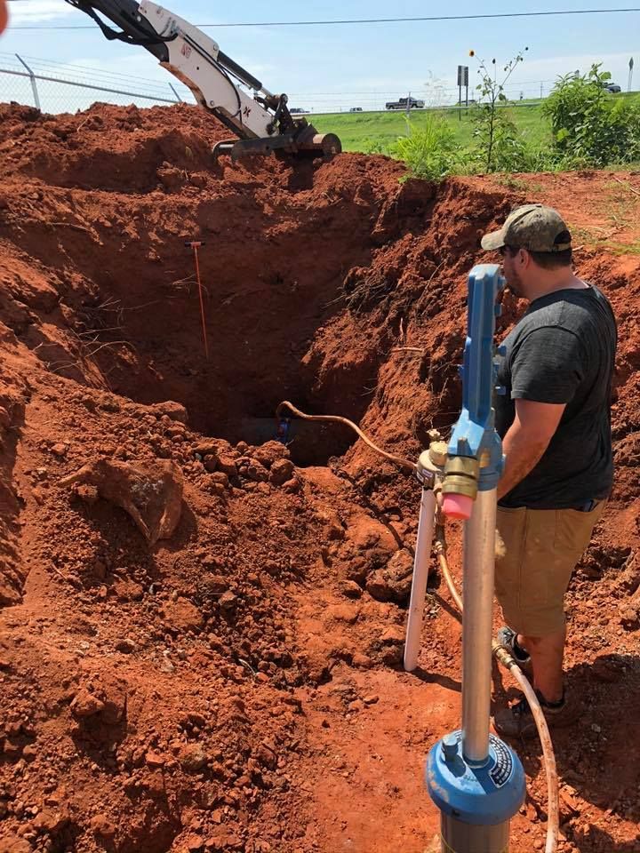 Man near well digging site. An excavator, red dirt, and blue well components are visible.