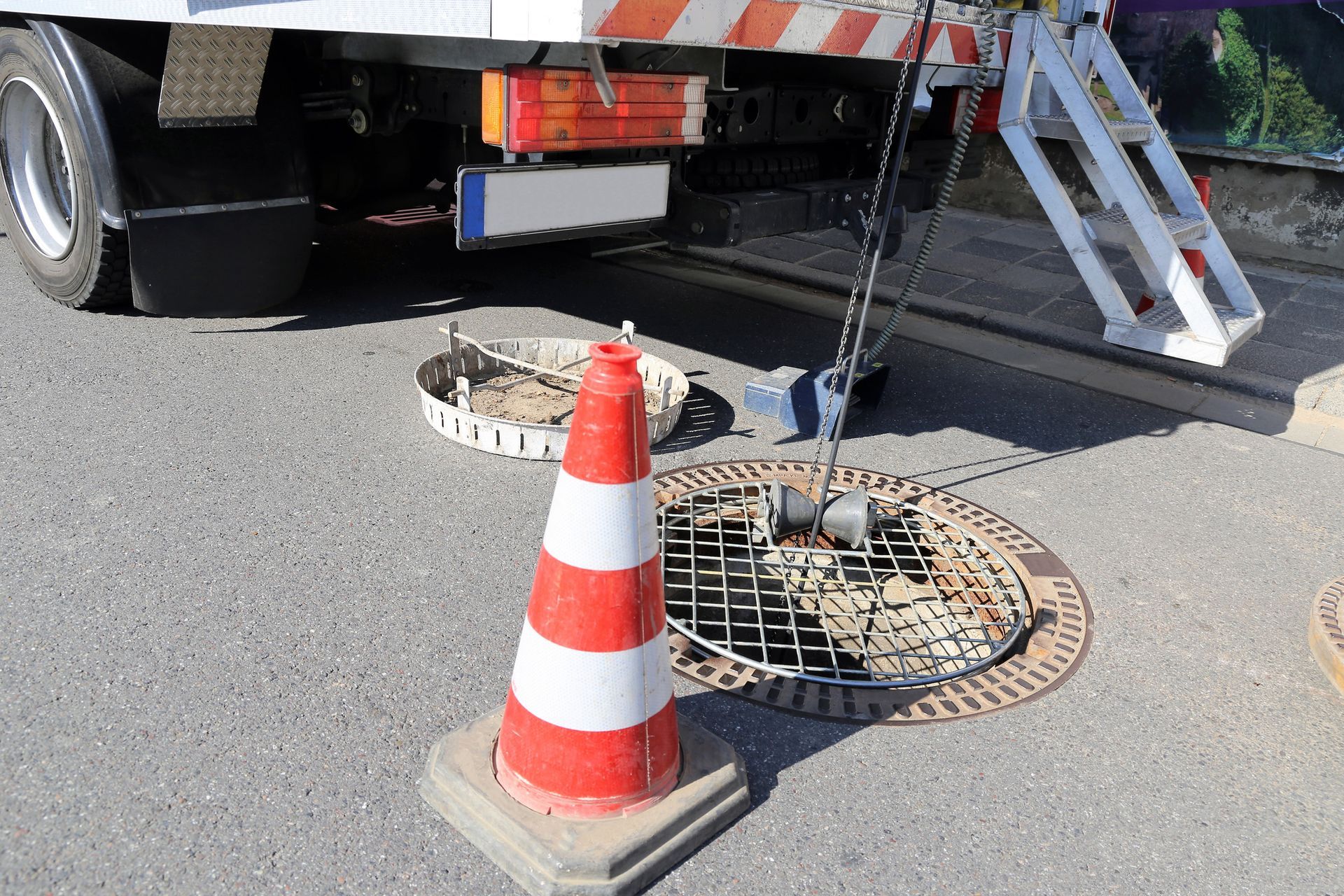 A road work scene: A truck near an open manhole, a safety cone in front.