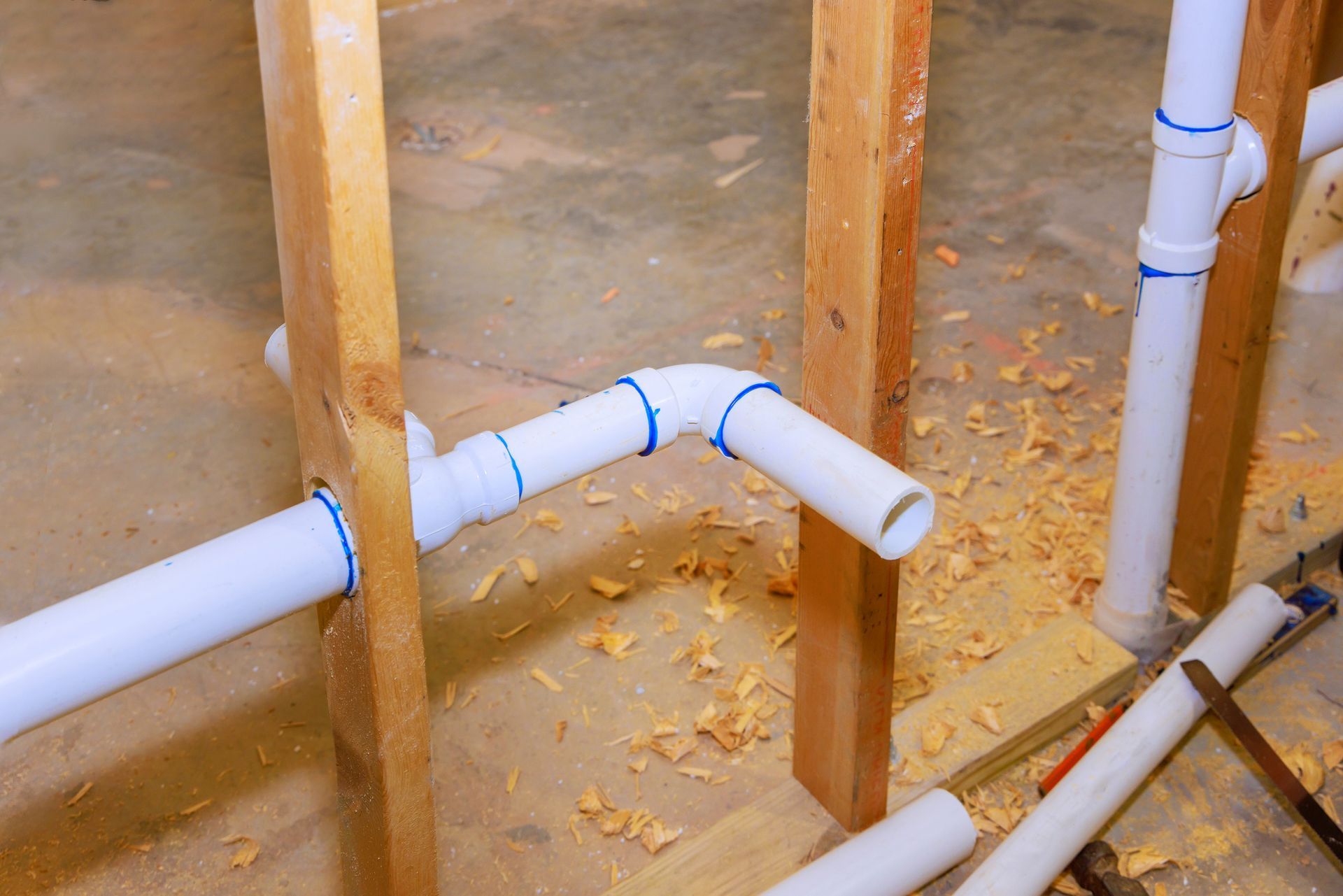 White PVC pipes with blue markings are installed between wooden studs; sawdust is present.