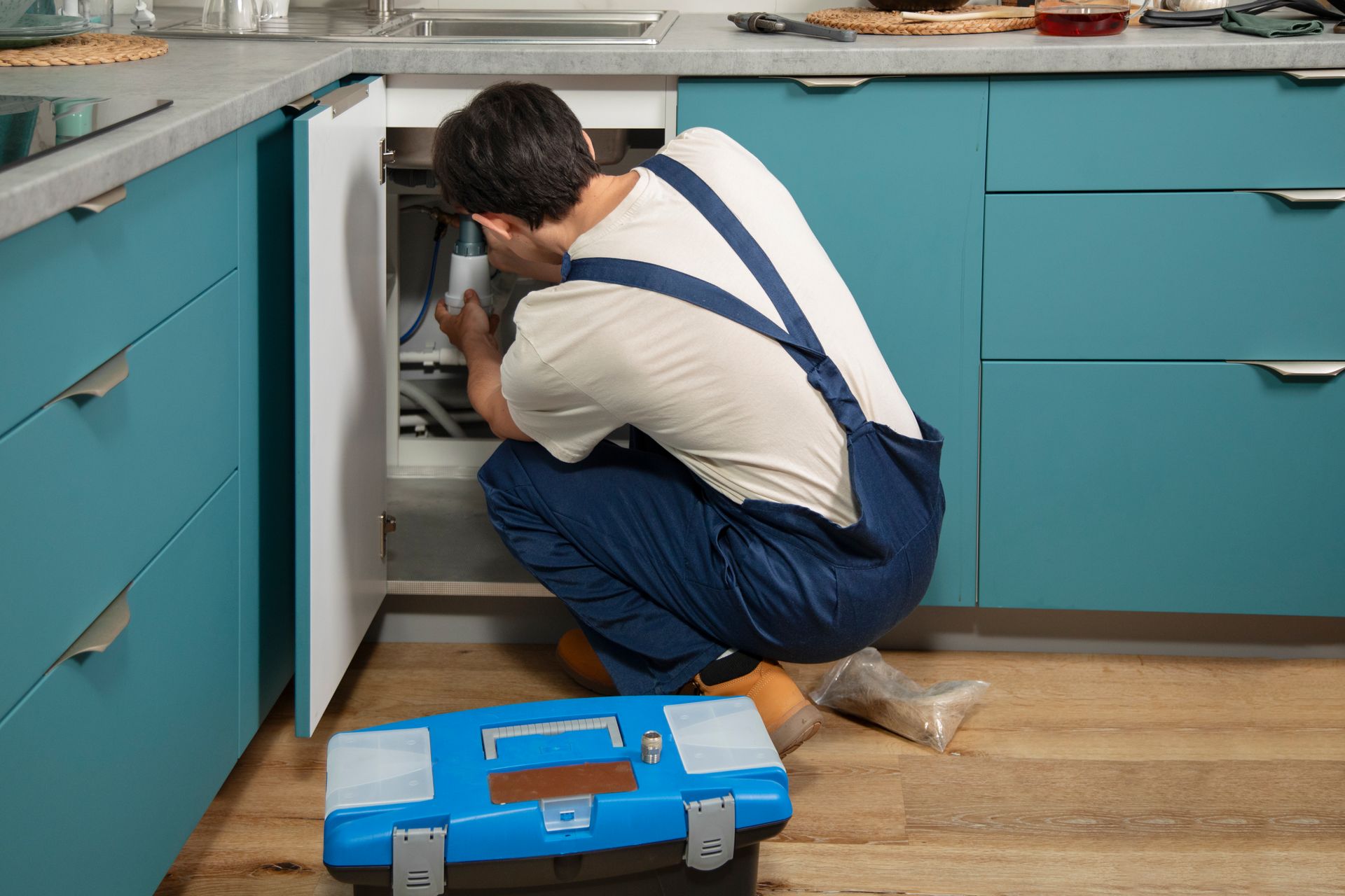 Man in overalls repairing appliance under blue kitchen cabinets. Blue toolbox on floor.