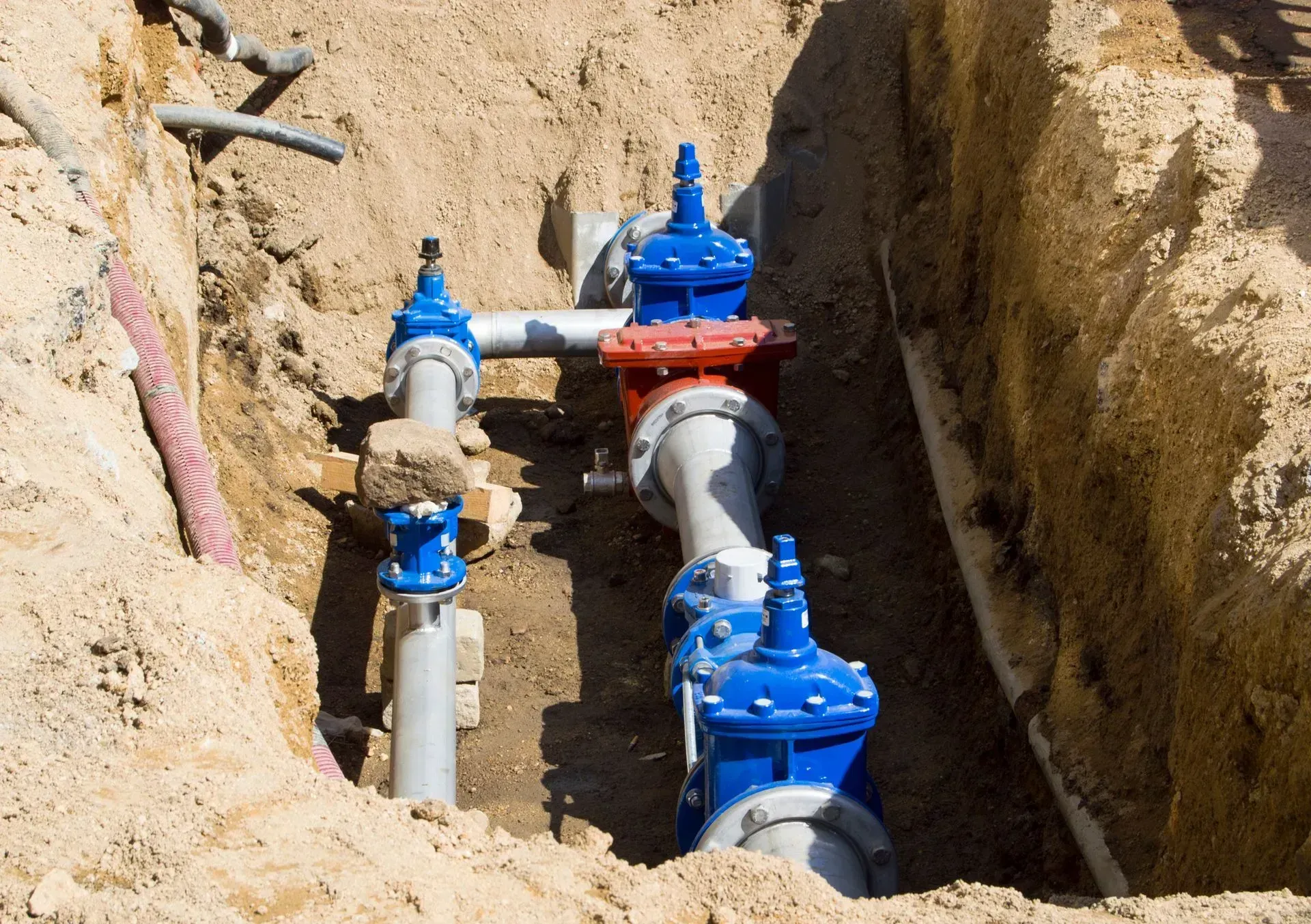 Pipes and valves in a trench, with blue and red components, likely for water or gas infrastructure.