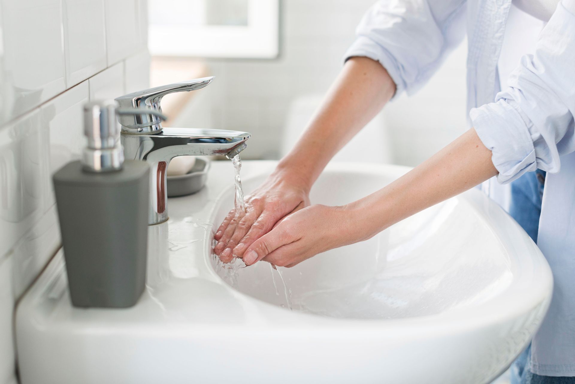 Person washing hands in a white bathroom sink under running water. A soap dispenser sits nearby.