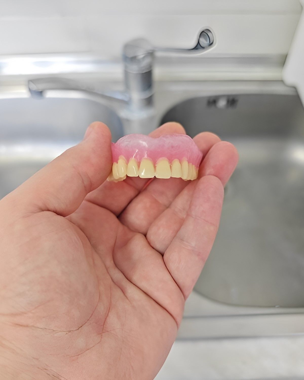 A Person is Holding a Denture in Their Hand in Front of a Sink — Yeppoon Denture Clinic in Rockhampton, QLD