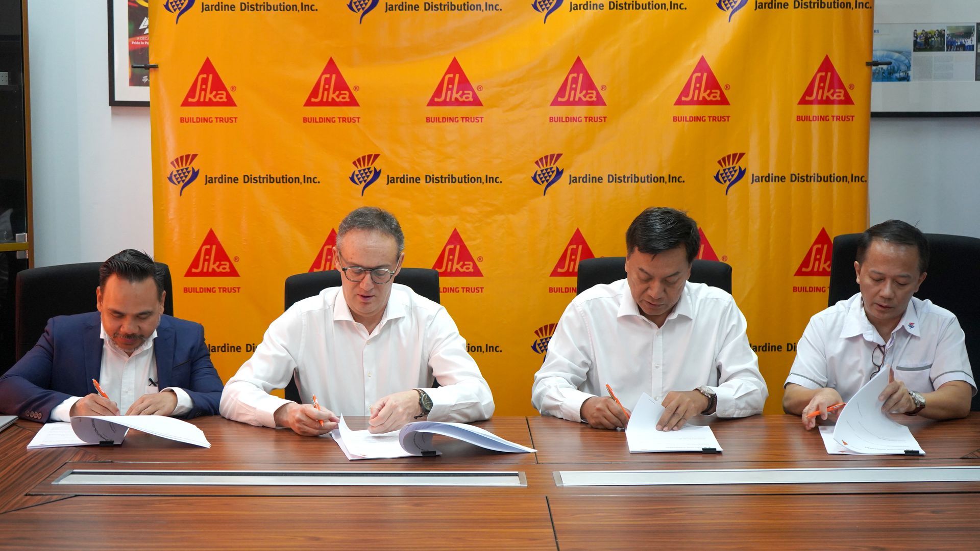 Four men signing documents at a table with a yellow Sika corporate backdrop.