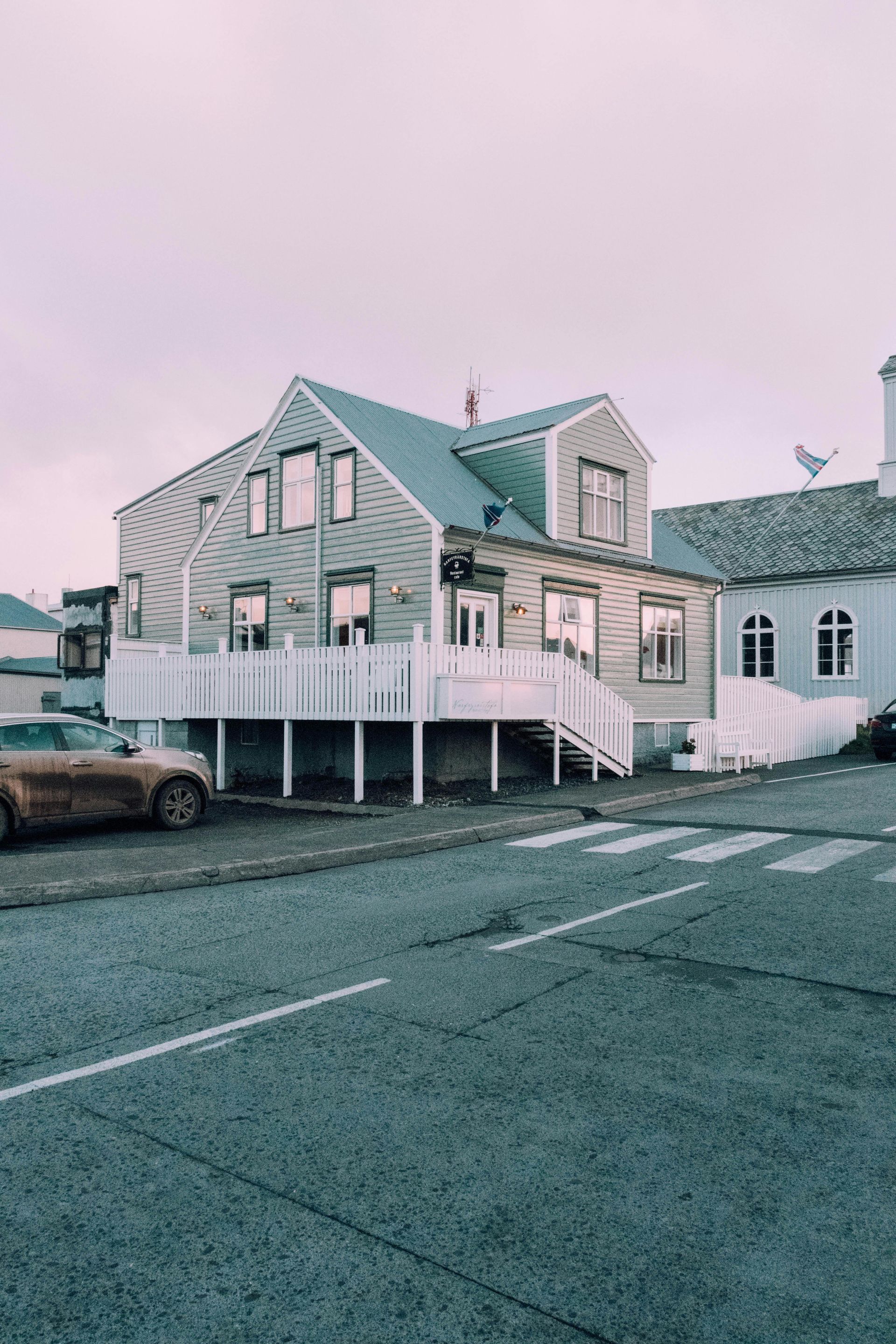Two-story house with a white deck, parked cars, and a road in a town. Overcast sky.