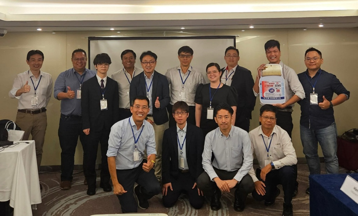 Group photo: A diverse group of people indoors, smiling, thumbs-up. Some kneeling, holding a box.