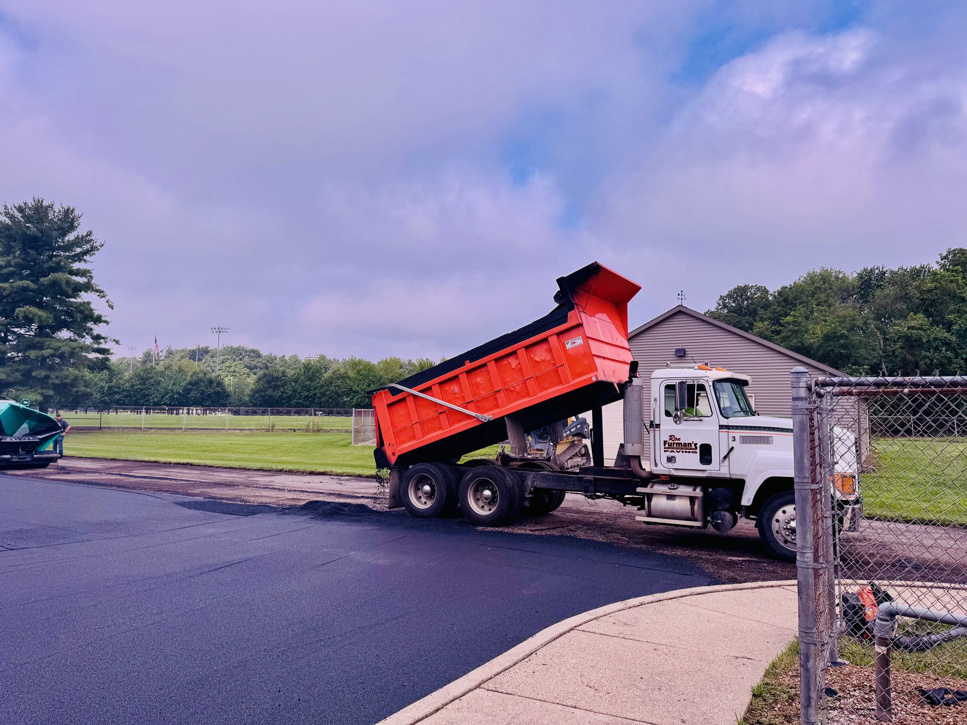 Orange dump truck unloading asphalt onto a newly paved surface, near a building and chain link fence.