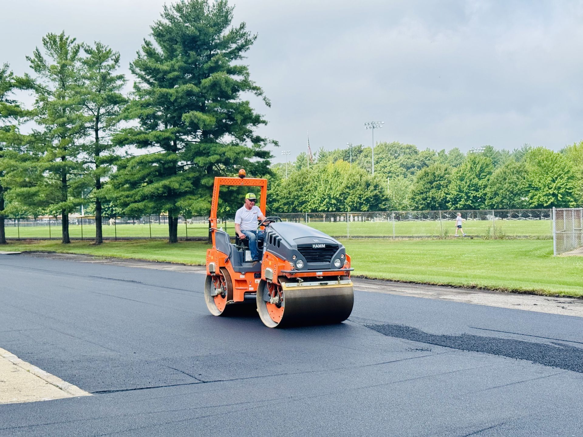 A man operating an orange asphalt roller on a newly paved road.