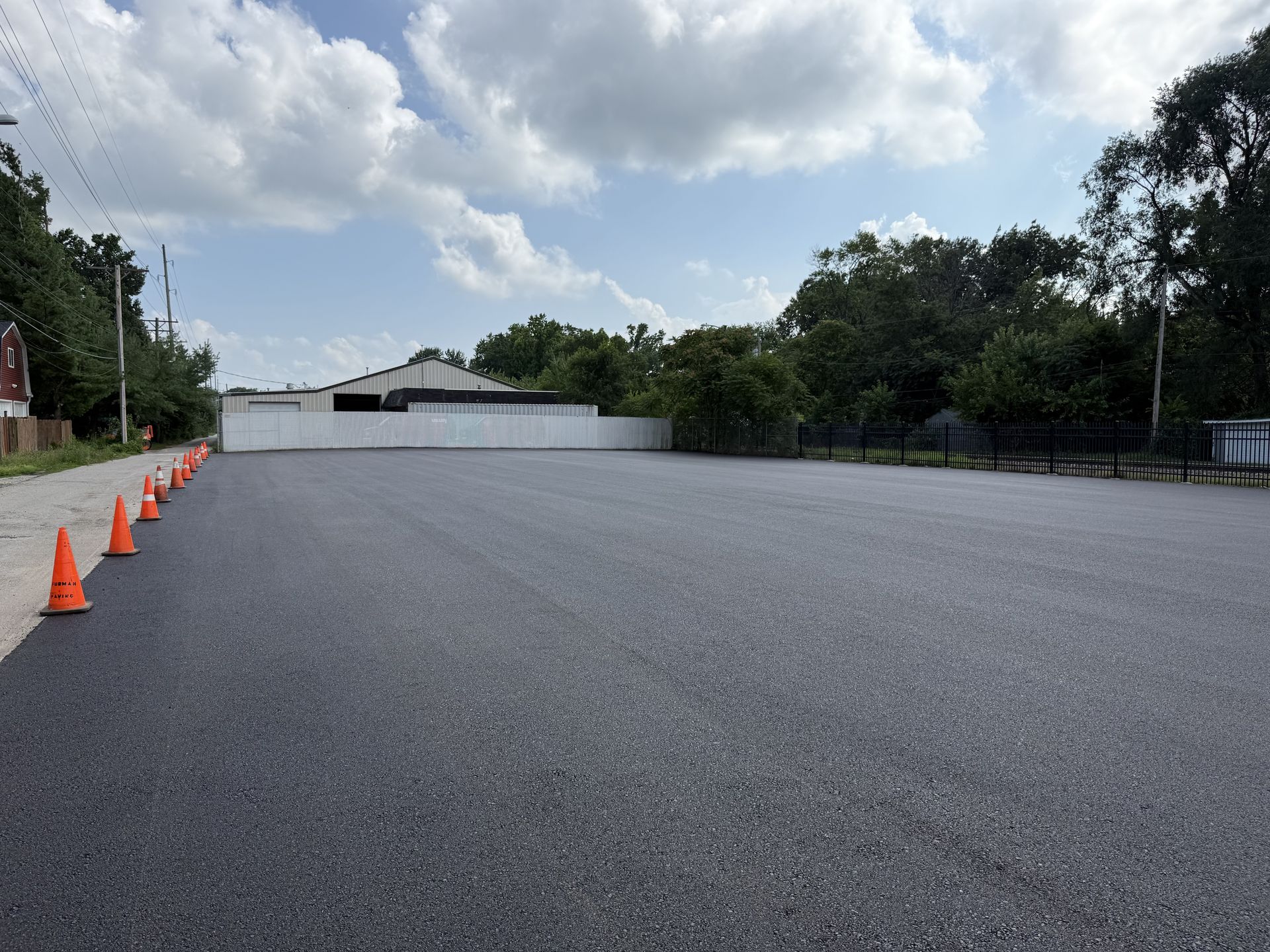 Newly paved asphalt lot with orange traffic cones, building in background, blue sky.