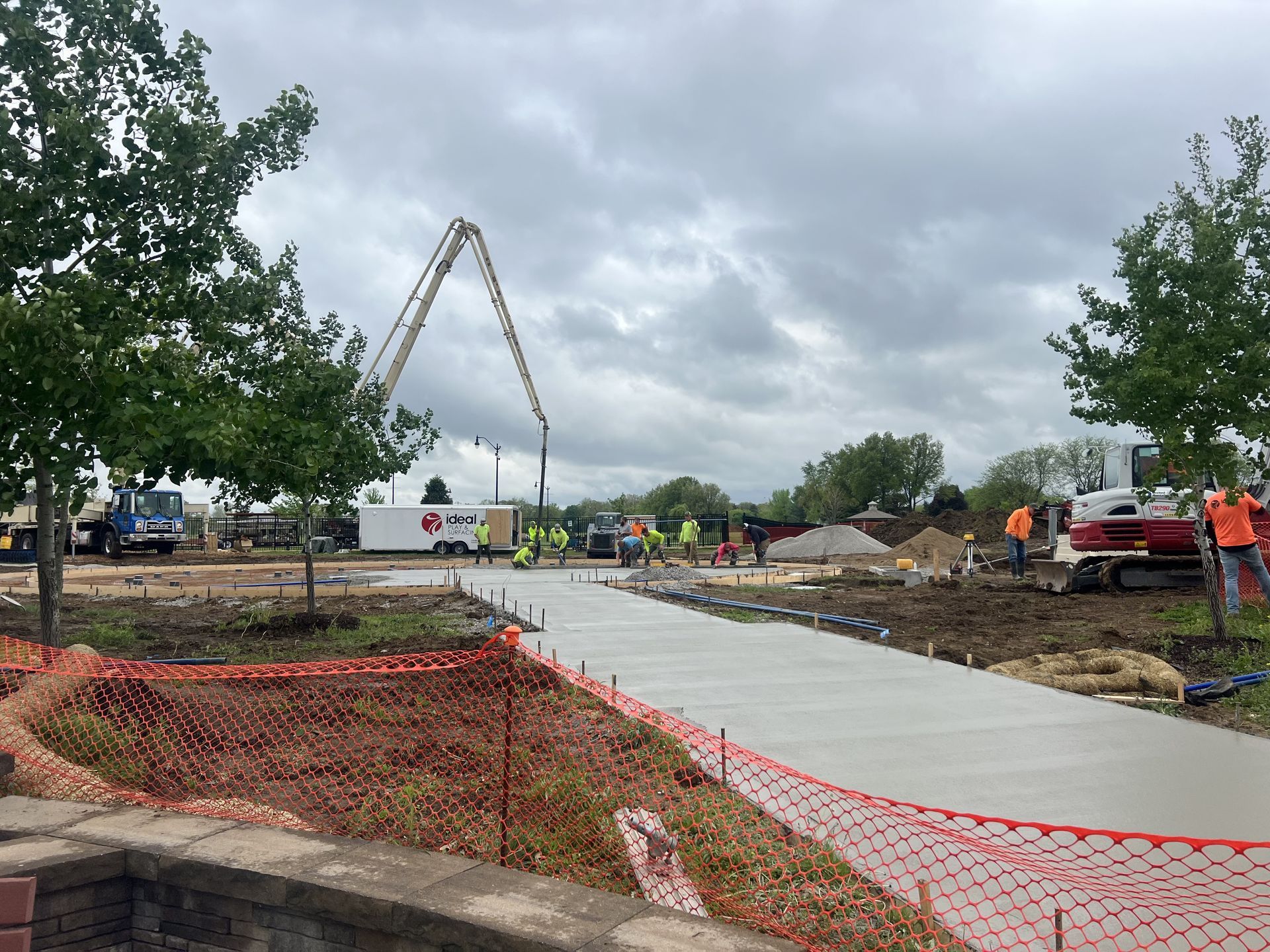 Construction site: concrete pouring for a pathway, orange safety fencing, workers, and a concrete pump truck under a cloudy sky.