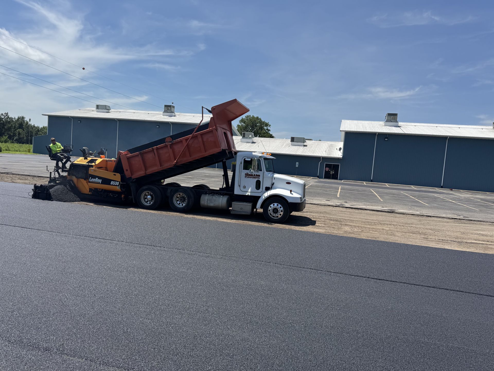 Asphalt paving: Dump truck unloading asphalt into a paving machine on a sunny day.