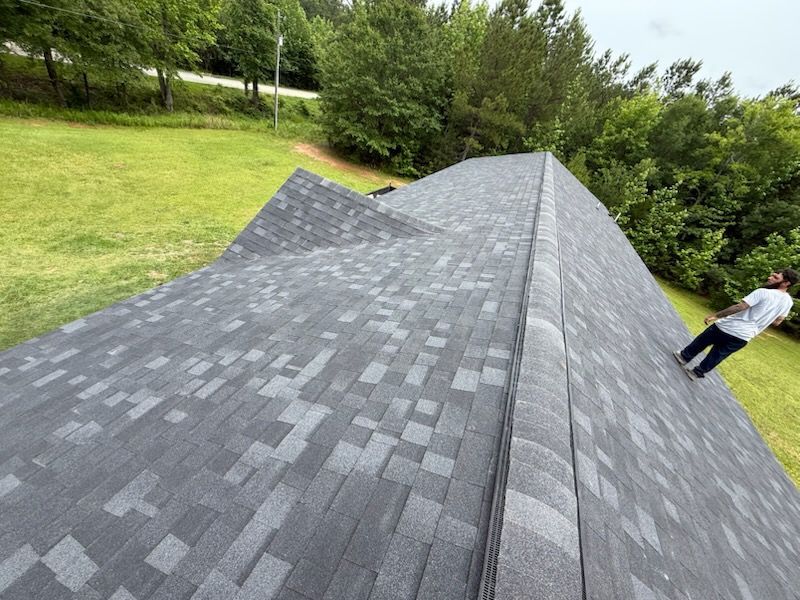 Man on a gray asphalt shingle roof, green lawn, trees in background.