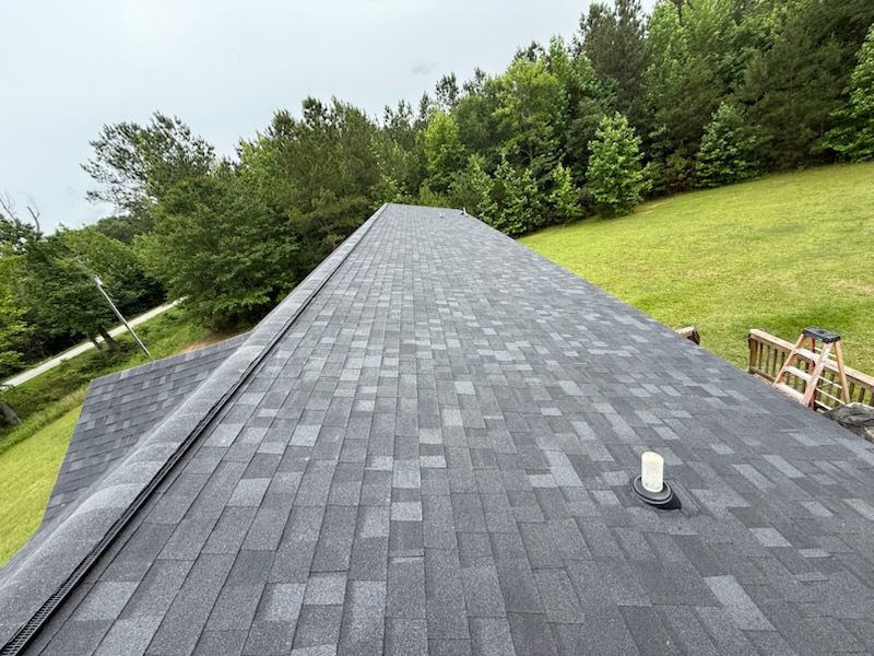 Dark gray shingle roof on a house, angled view, with trees and grass in the background.