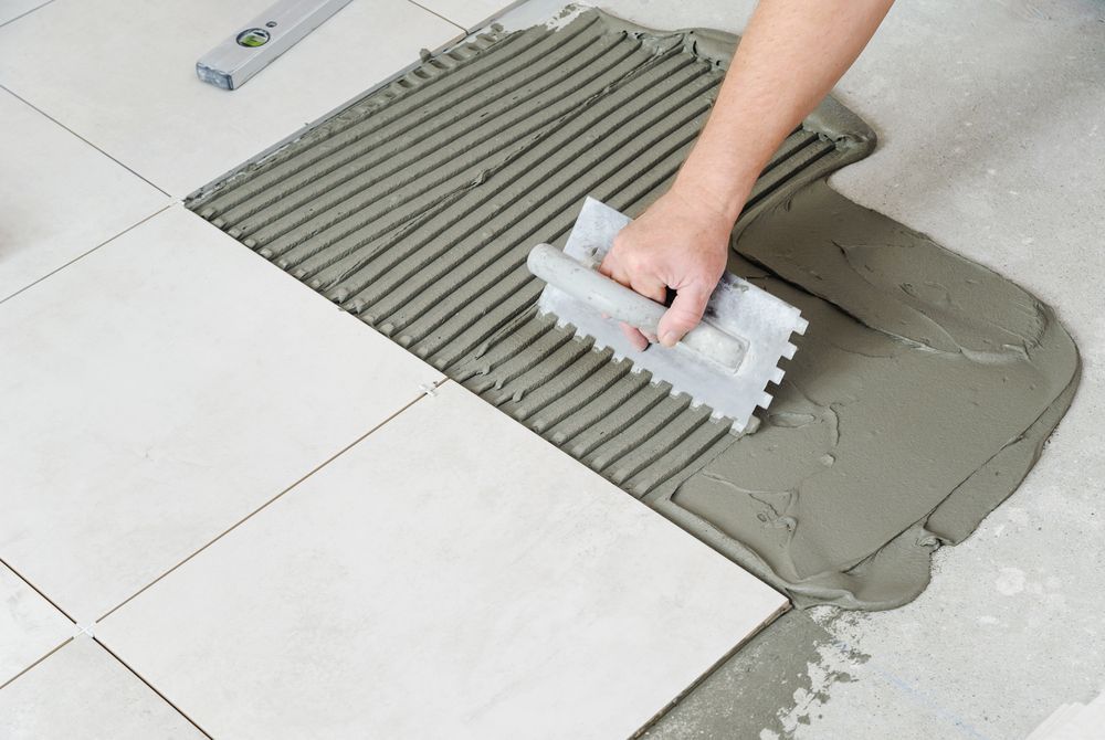 Person tiling a floor, applying mortar with a notched trowel to lay square tiles.