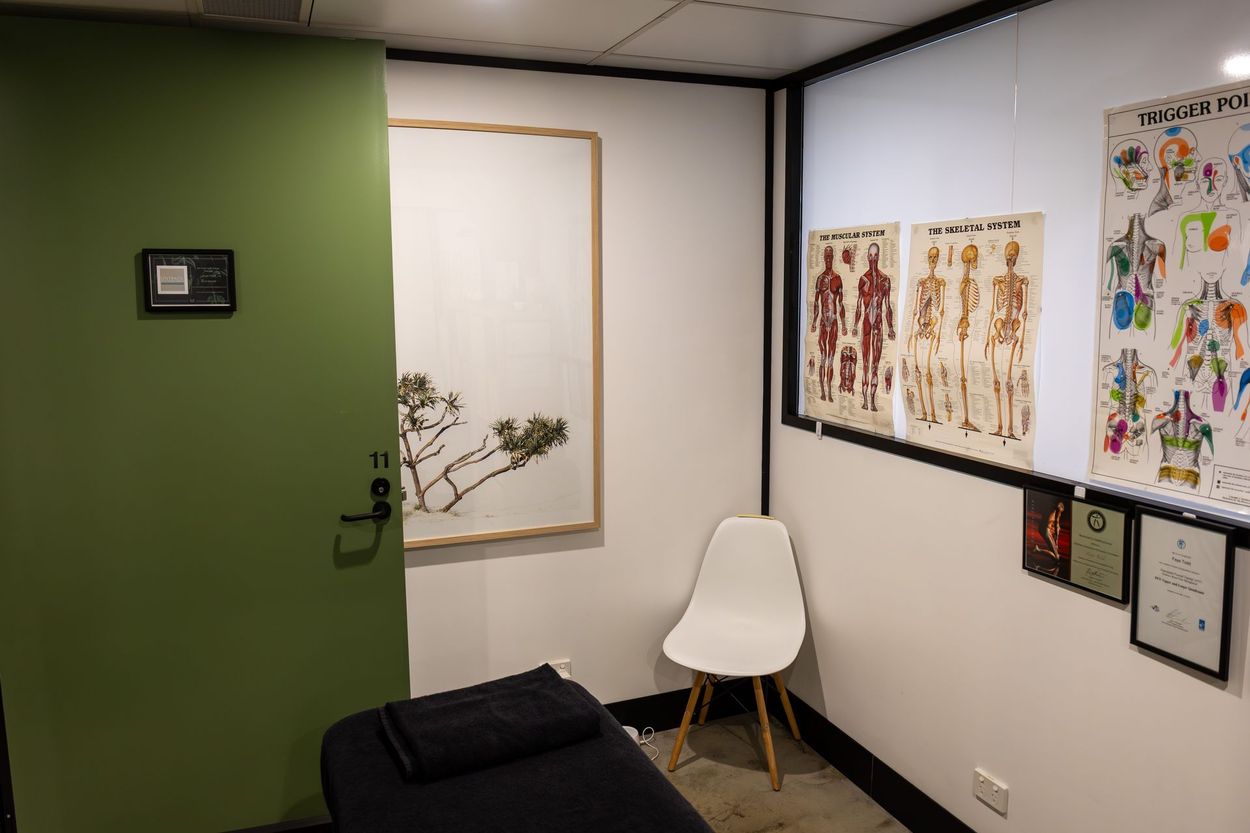 A treatment room with a massage table, chair, and anatomical charts. Green door on left.