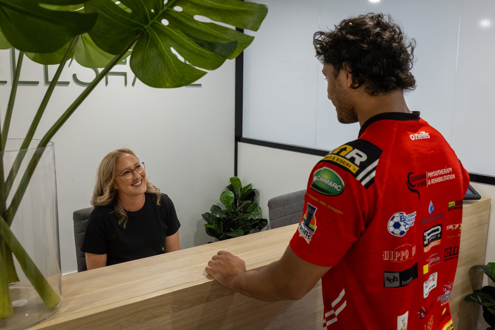 Man in red jersey at reception desk, speaking to woman behind the desk. Office setting, plants visible.