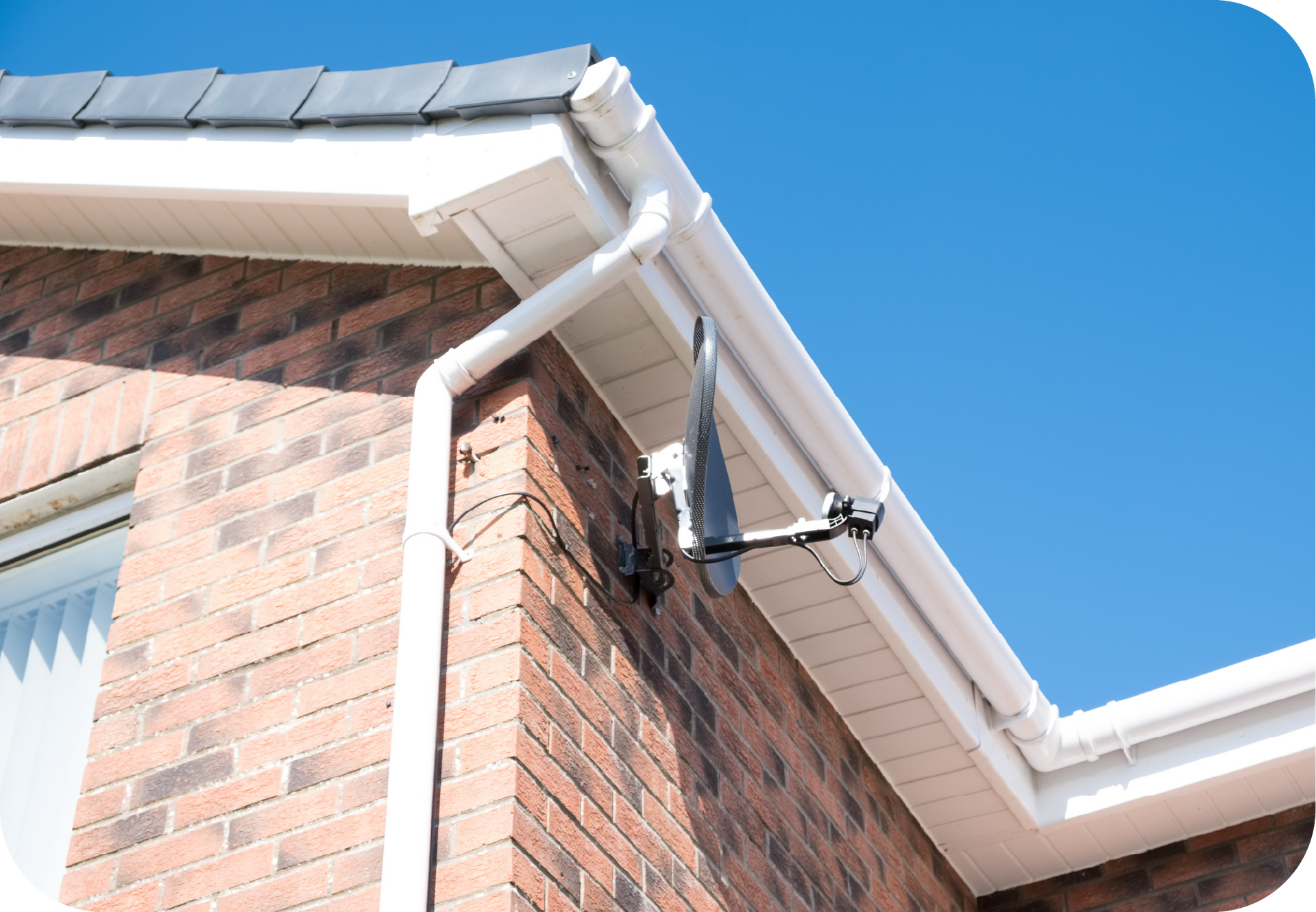 White gutter and downspout on a brick building with a mounted black antenna. Clear blue sky.