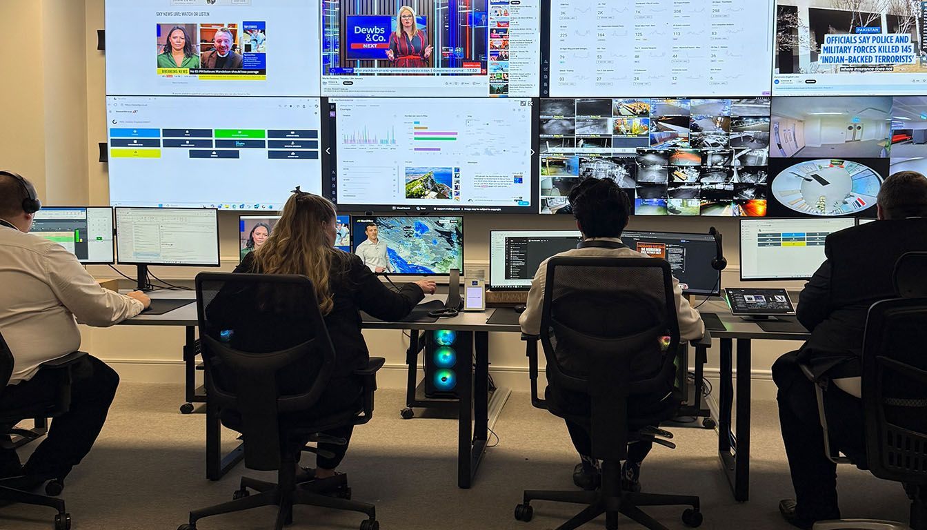 Four people working at desks in a control room with multiple screens displaying data and media.