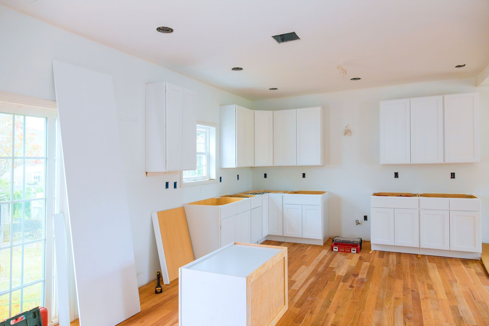 Bright kitchen under renovation with white cabinets, wood floors, and scattered tools on the floor