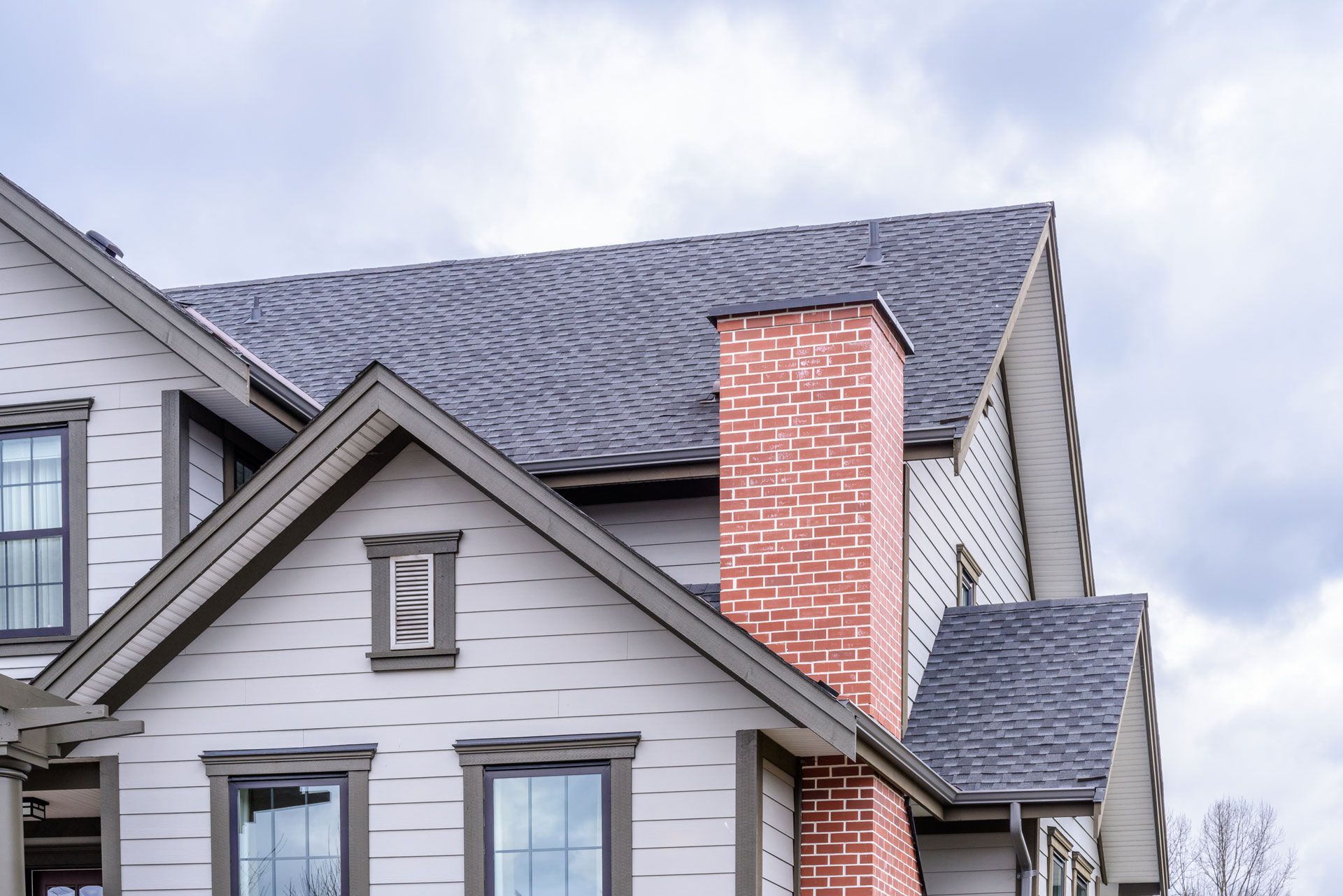 Close-up of a house roof and dormers with gray siding and a red brick chimney under a cloudy sky