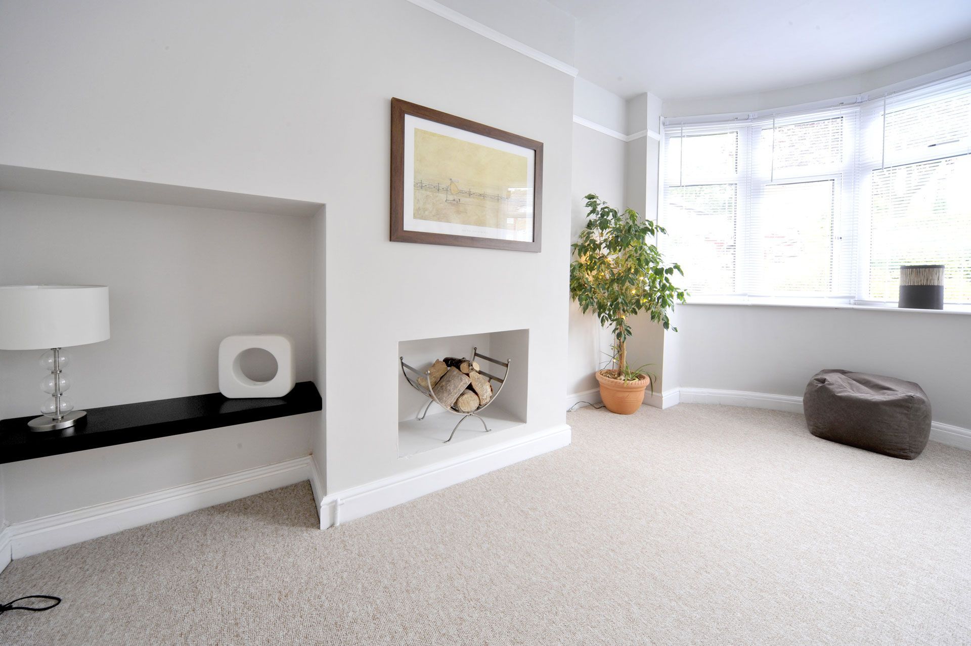 Bright living room with a built-in alcove, fireplace, bay window, potted plant, and gray ottoman