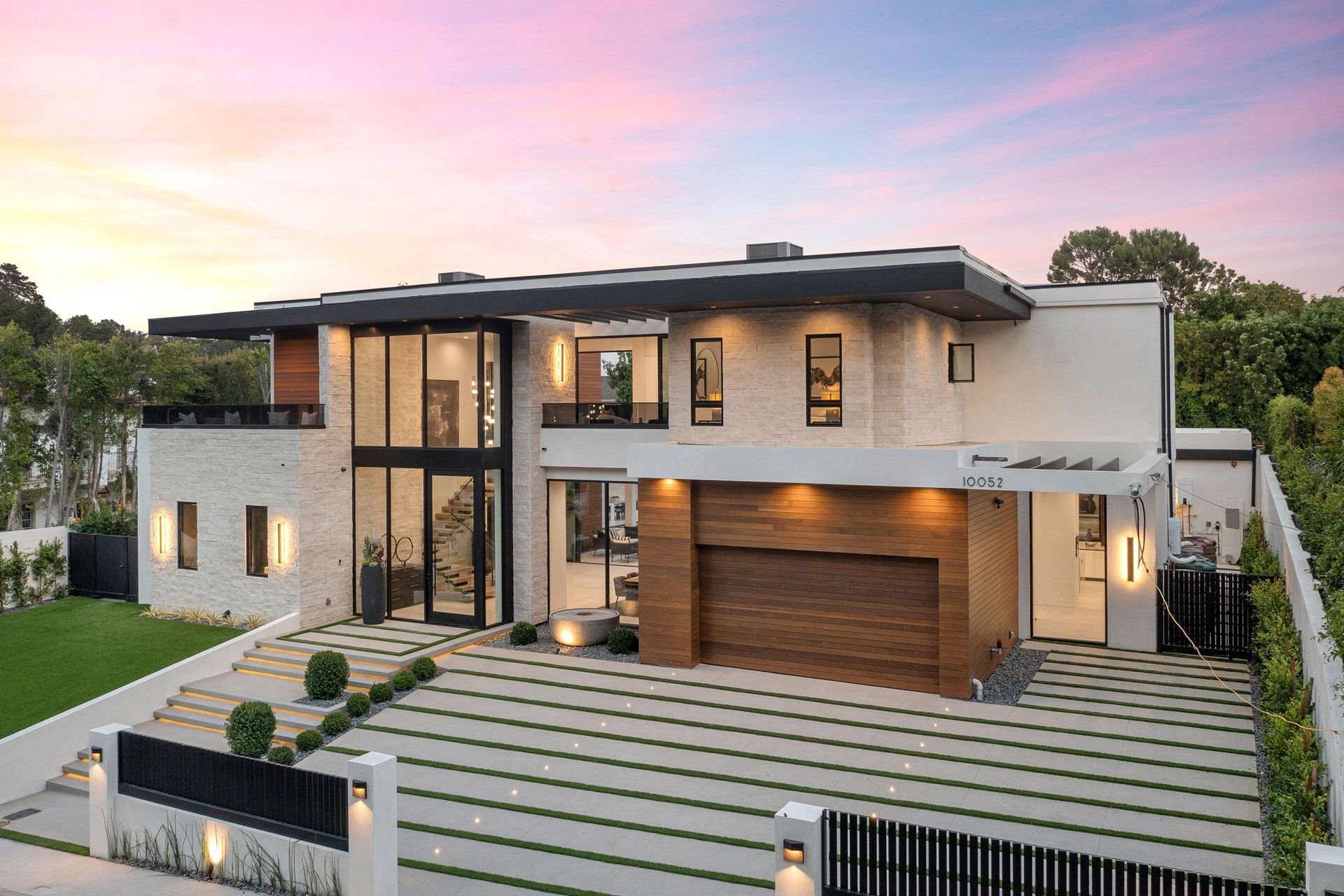 Modern two-story luxury house with a wood garage, glass entry, and landscaped front yard at sunset