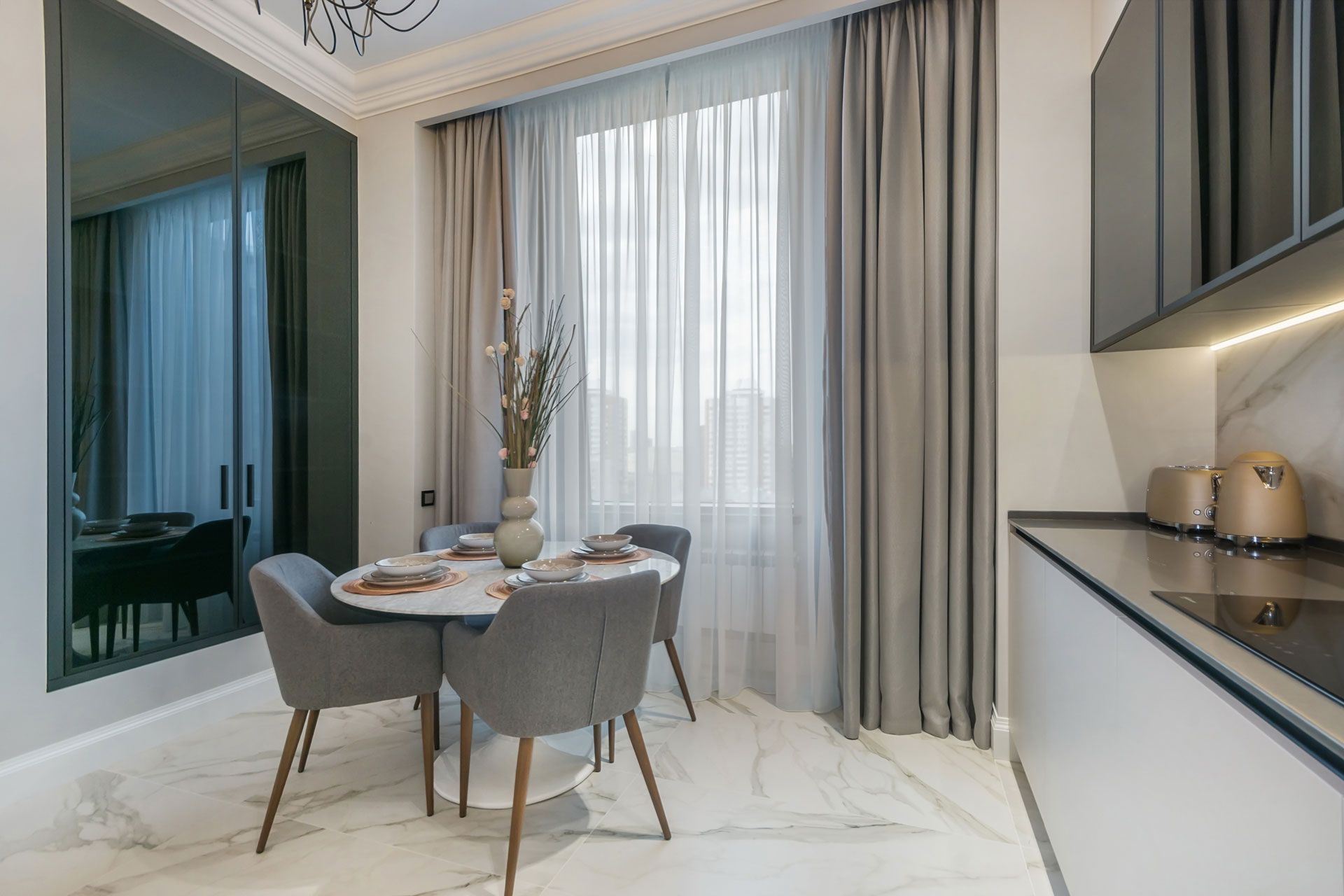 Modern dining area with gray chairs, a round table, white curtains, and a sleek kitchen counter