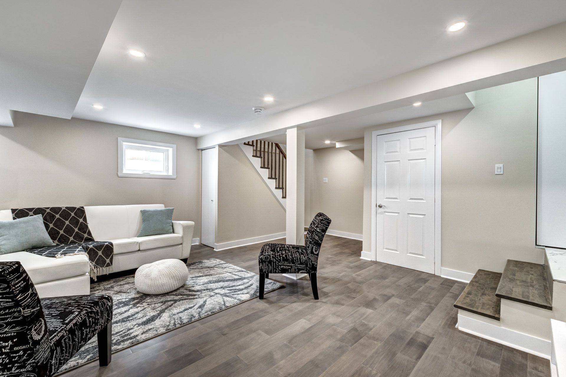Bright finished basement living room with gray sofa, patterned chairs, staircase, and white closet doors.