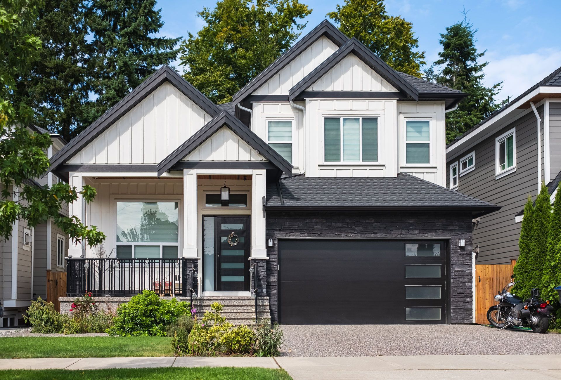 Modern two-story gray house with black garage door and front porch, surrounded by trees and shrubs