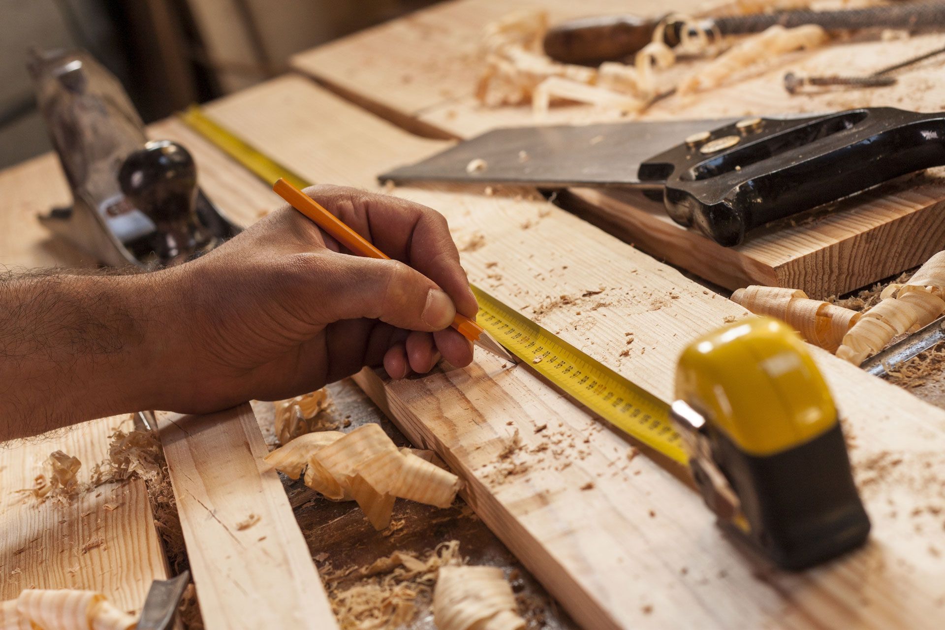 Hand using a pencil and tape measure on a wooden board in a workshop, with tools nearby