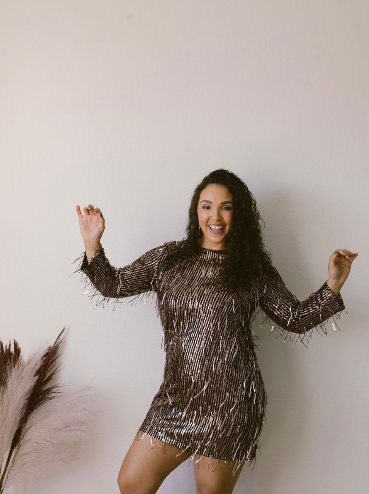 A woman in a sequined dress is dancing in front of a white wall.