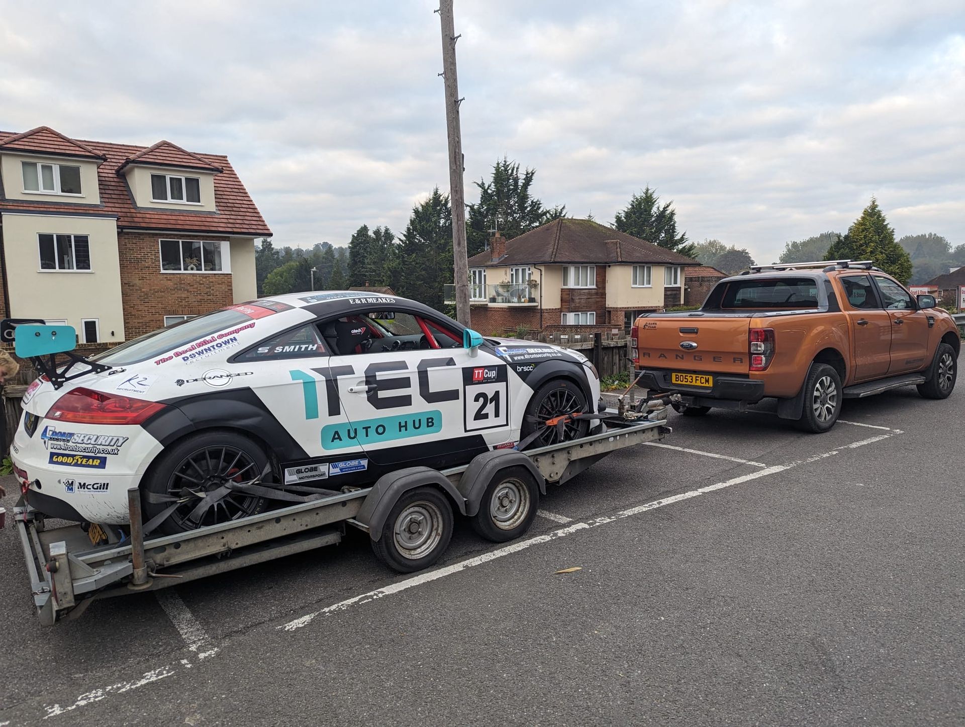 Orange truck towing a race car on a trailer in a parking lot. Buildings in background.
