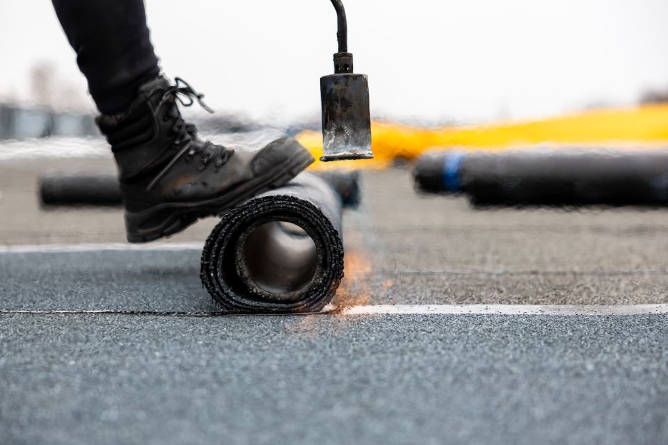 Roofer torching a rolled roofing membrane on a flat roof. Black boot, flame, and asphalt surface.