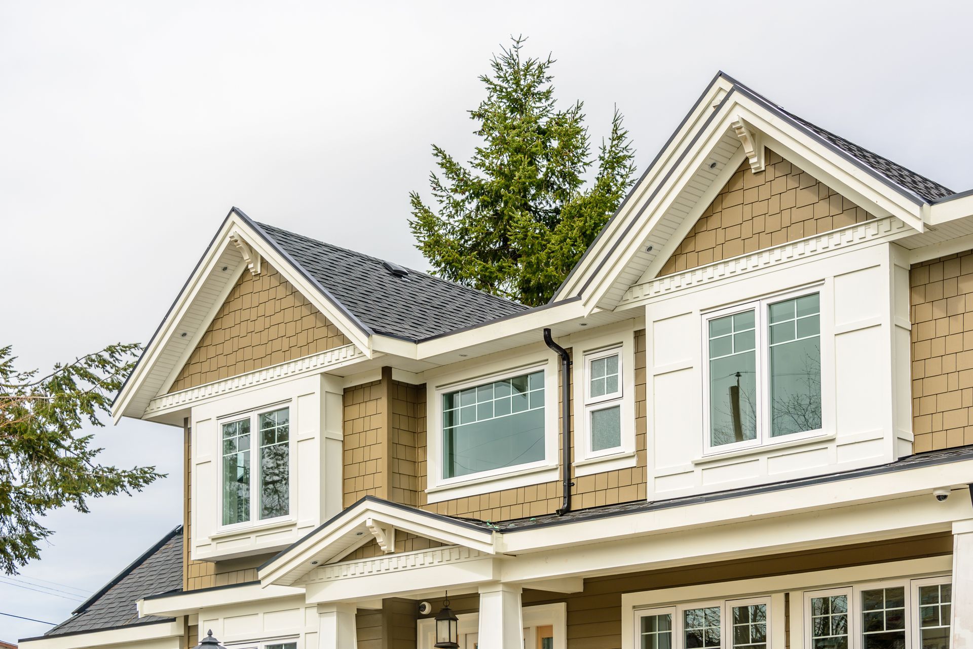 Two-story house with tan siding, white trim, and dark gray roof. A tree is visible in the background.