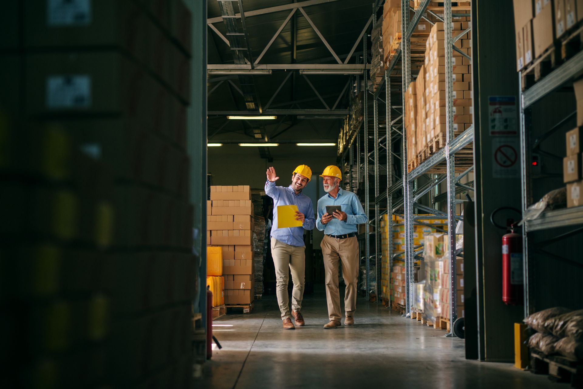 Two workers in a warehouse aisle, wearing yellow hard hats.