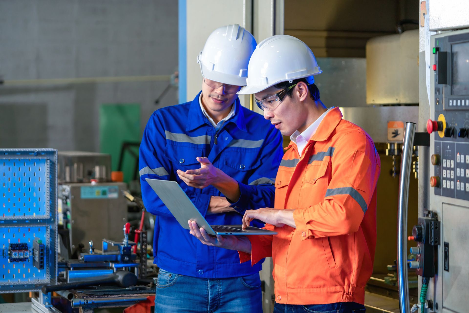 Two engineers in workwear and hard hats consult a laptop in a factory setting.